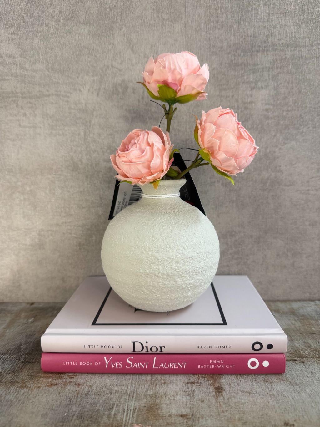 Vakkenkast decoratieset met beige bolle vaas, drie roze kunstpioenrozen en twee roze/witte tafelboeken over Dior en Yves Saint Laurent op houten ondergrond.