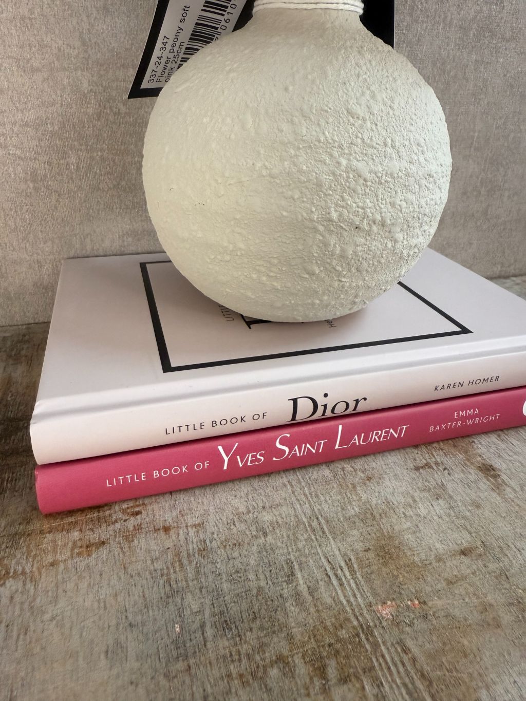 Vakkenkast decoratieset met beige bolle vaas, drie roze kunstpioenrozen en twee roze/witte tafelboeken over Dior en Yves Saint Laurent op houten ondergrond.