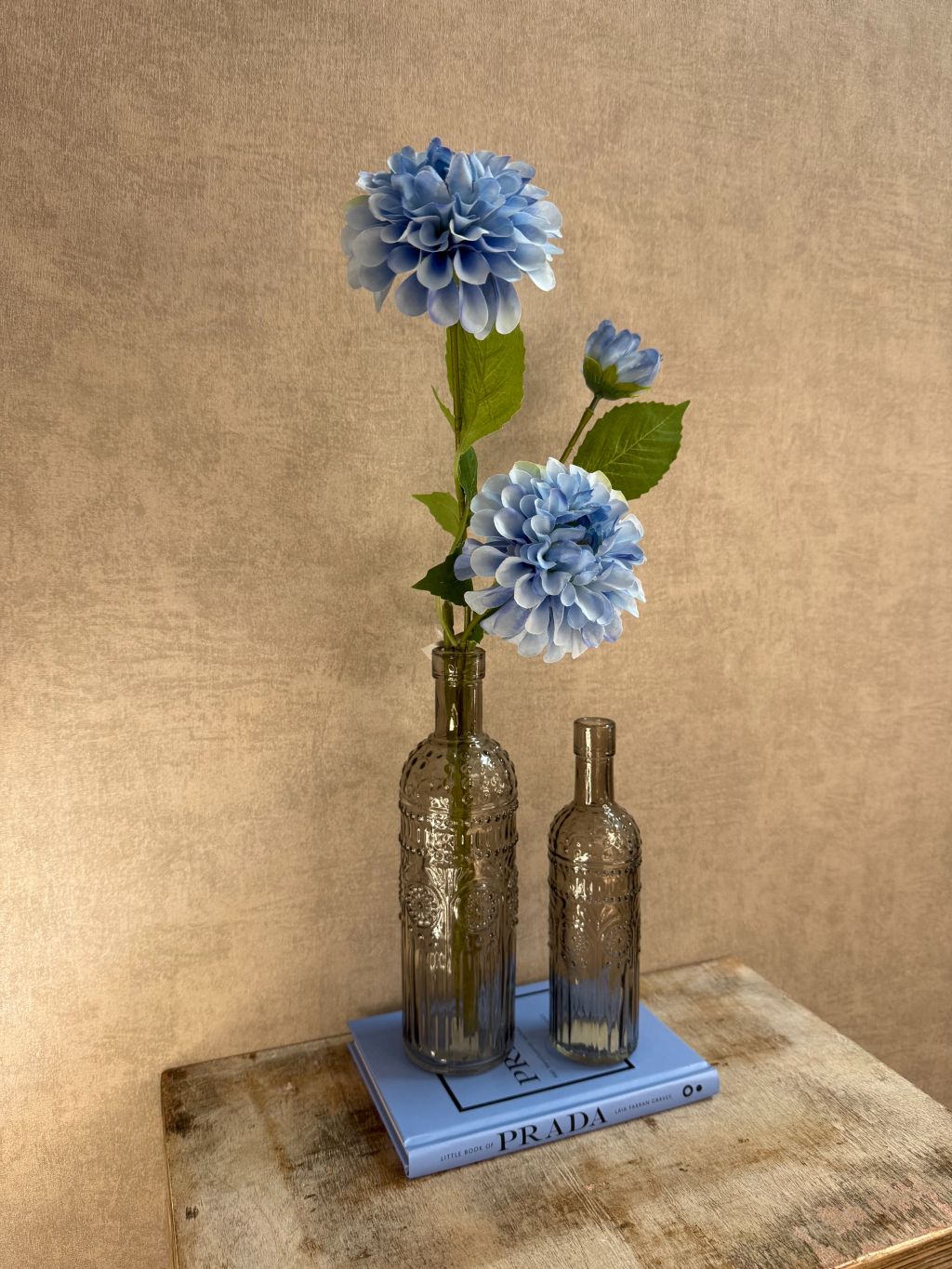 Decoratieset met lichtblauw Prada-boek, twee rookglazen flesvazen en blauwe kunst dahlia tegen een warme beige achtergrond.