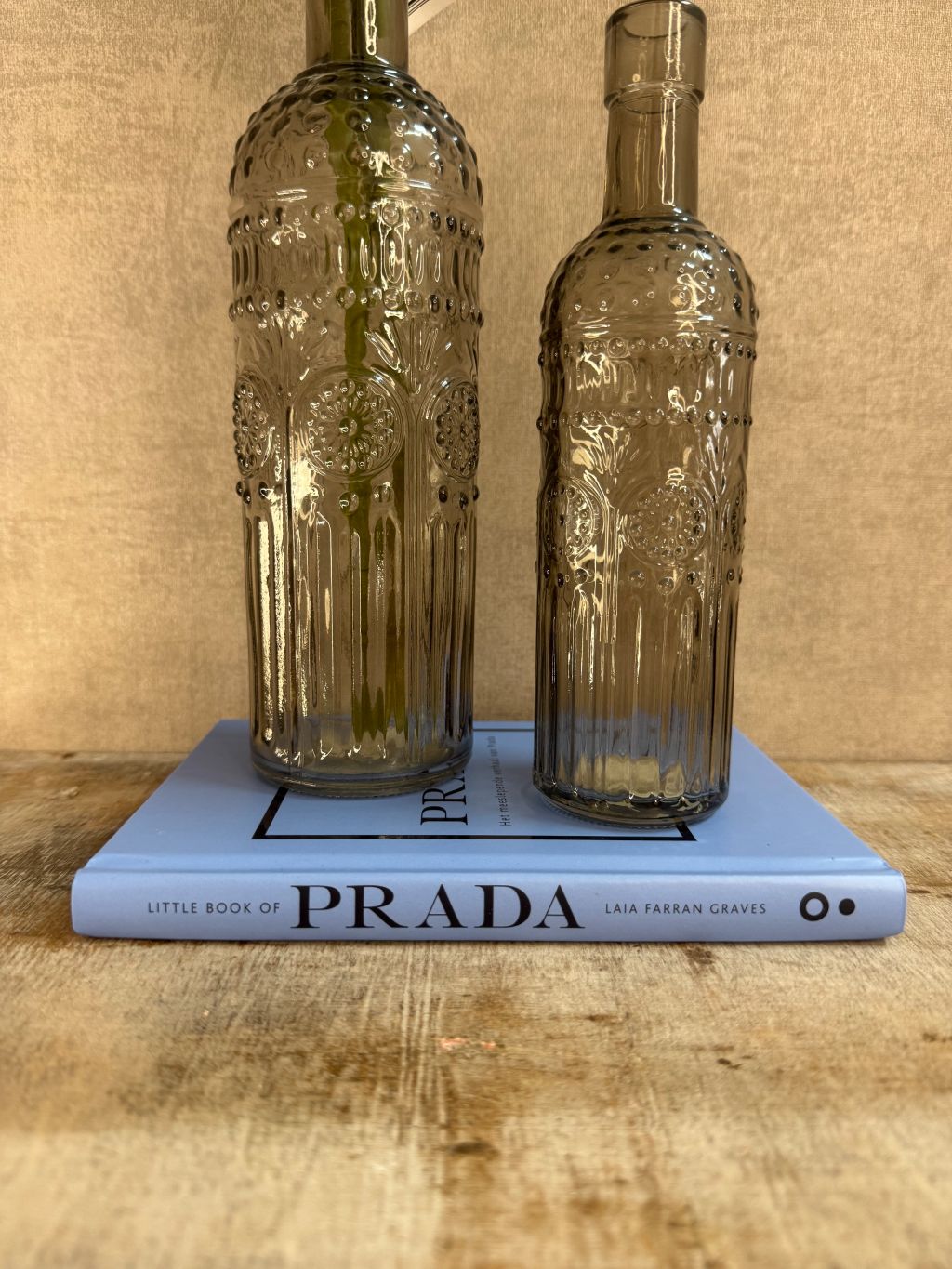 Decoratieset met lichtblauw Prada-boek, twee rookglazen flesvazen en blauwe kunst dahlia tegen een warme beige achtergrond.