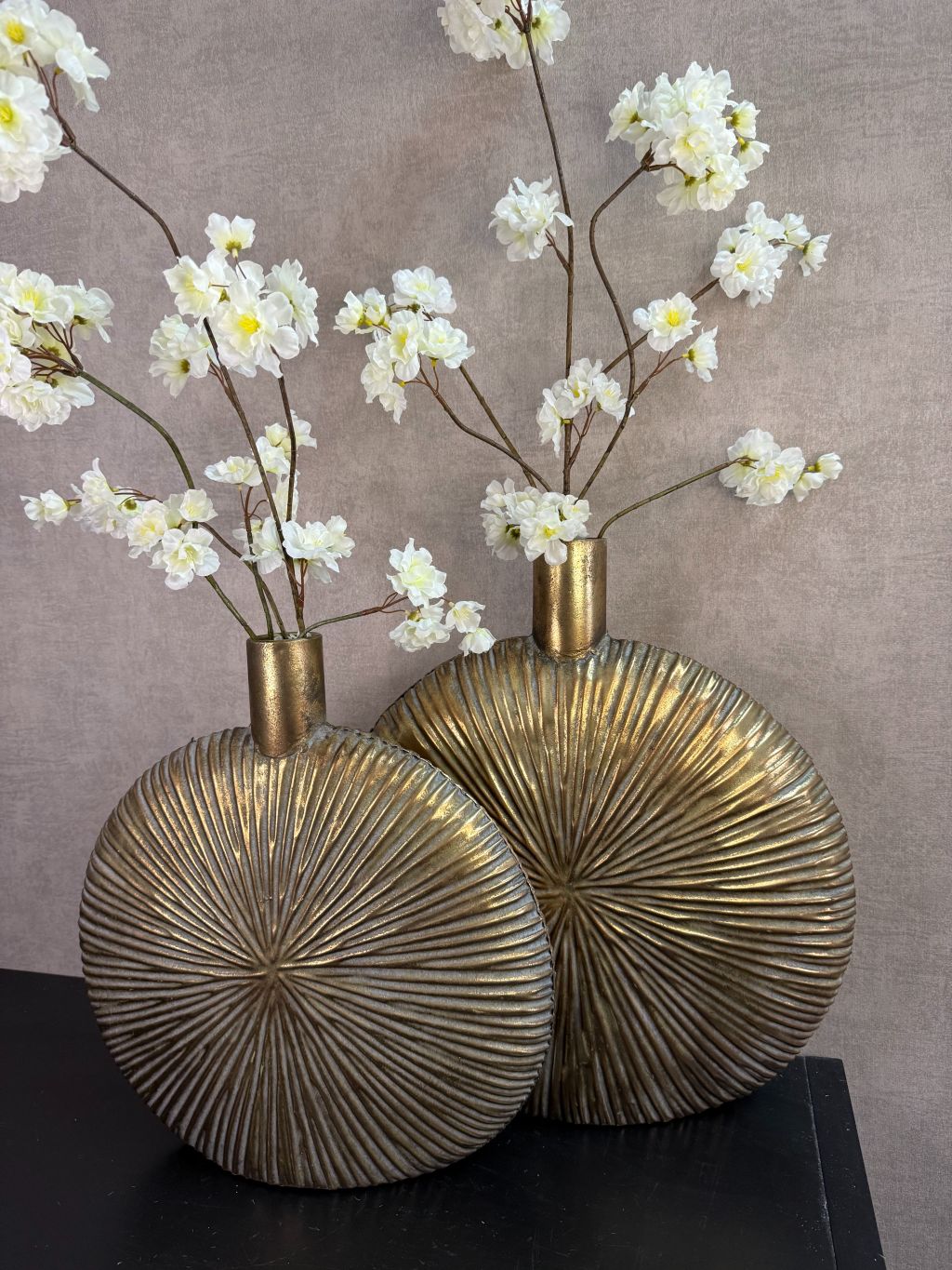 Twee goudkleurige decoratieve vazen met witte bloesemtakken op een zwarte ondergrond tegen een neutrale achtergrond.