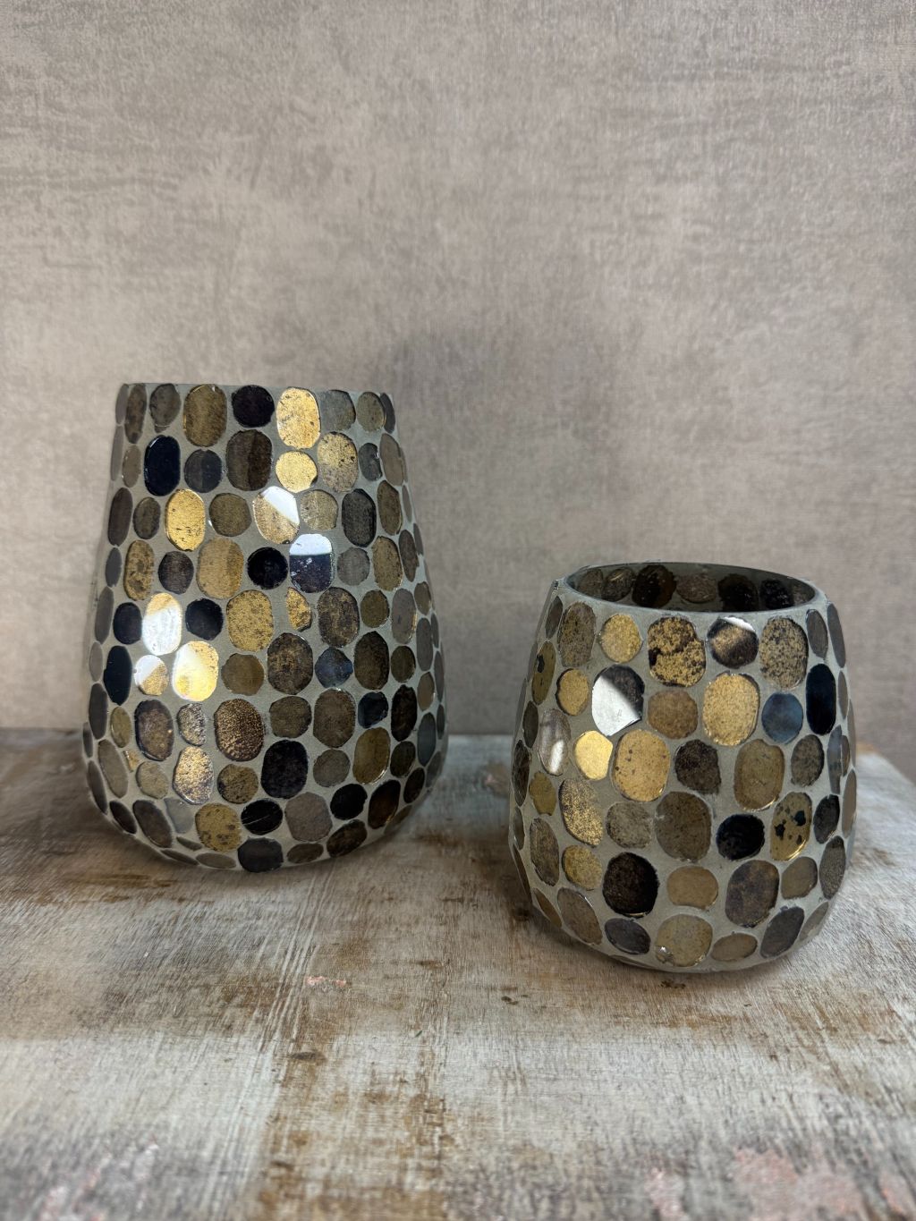 Twee glazen windlichten met mozaïek in aardse tinten van bruin, goud en zwart, staand op een houten ondergrond tegen een grijze muur.