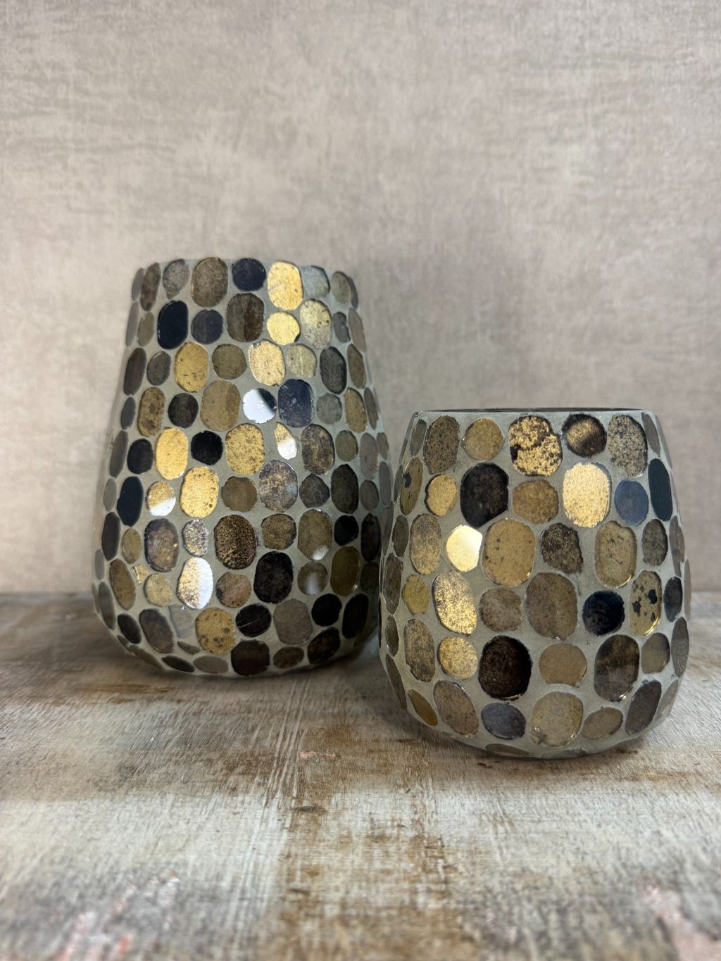 Twee glazen windlichten met mozaïek in aardse tinten van bruin, goud en zwart, staand op een houten ondergrond tegen een grijze muur.