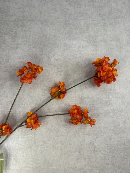 Zijden bloesemtak met oranje bloemen liggend op een houten oppervlak, met levendige bloesems en subtiel groene steel.