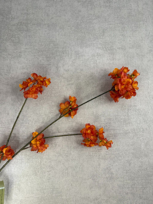 Zijden bloesemtak met oranje bloemen liggend op een houten oppervlak, met levendige bloesems en subtiel groene steel.