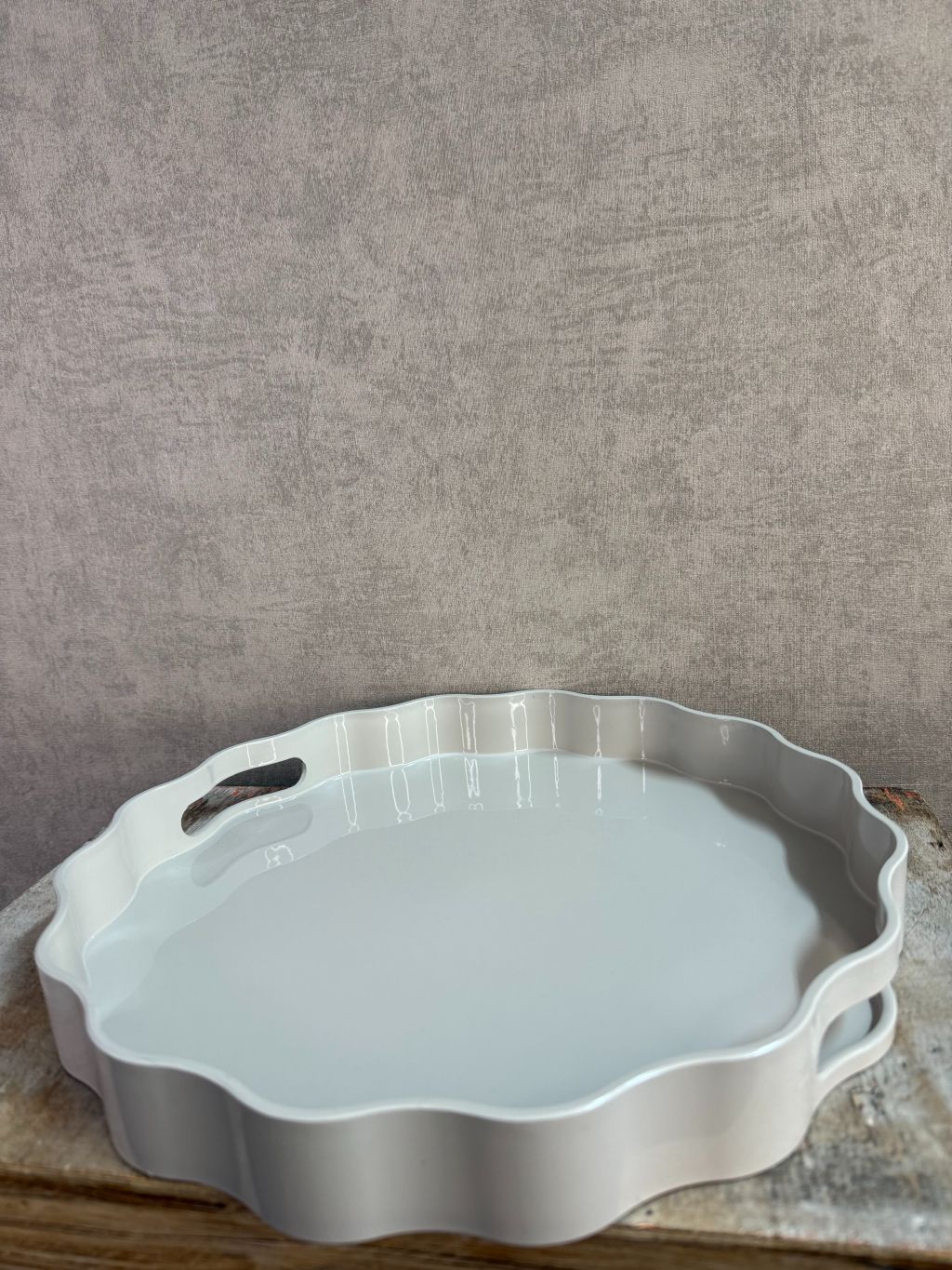 Rond dienblad in grijs-beige kleur met golvende rand en handgrepen, geplaatst op een houten ondergrond.