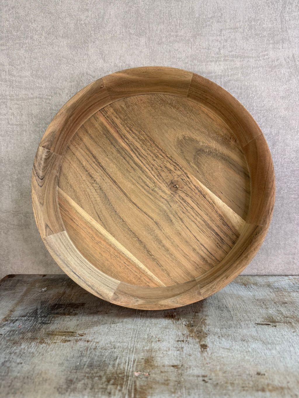 Rond dienblad van licht acaciahout met zichtbare houtnerf, geplaatst op een houten ondergrond tegen een neutrale achtergrond.
