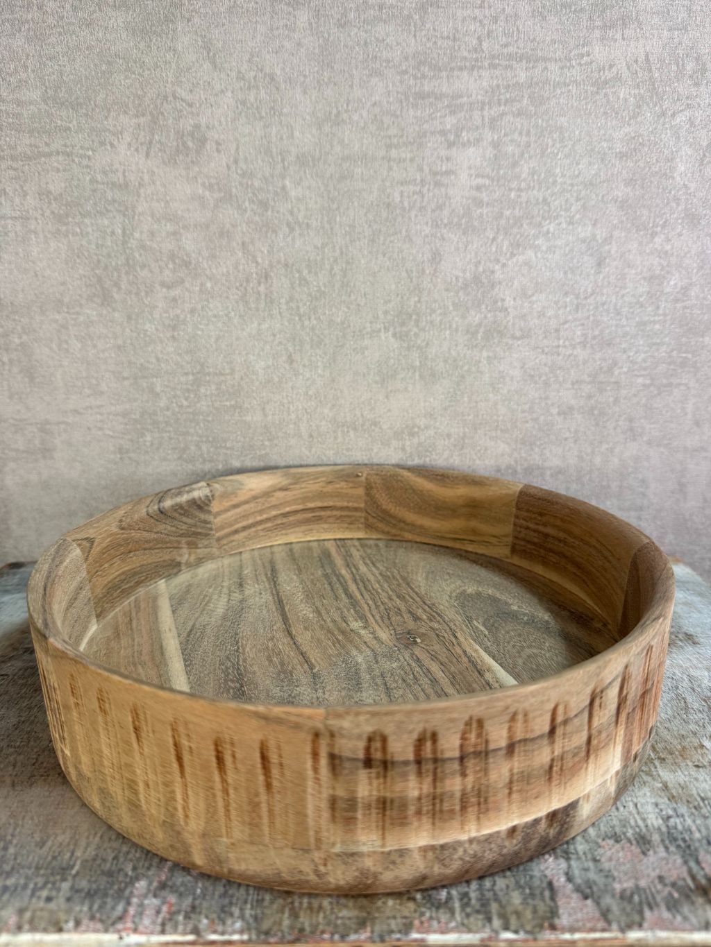 Rond dienblad van licht acaciahout met zichtbare houtnerf, geplaatst op een houten ondergrond tegen een neutrale achtergrond.