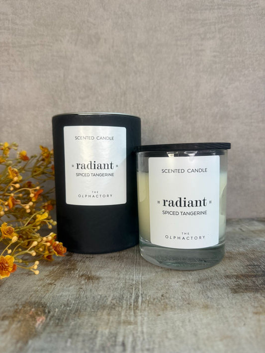 Geurkaars Radiant Spiced Tangerine van The Olphactory in glas met zwarte deksel, op houten ondergrond.