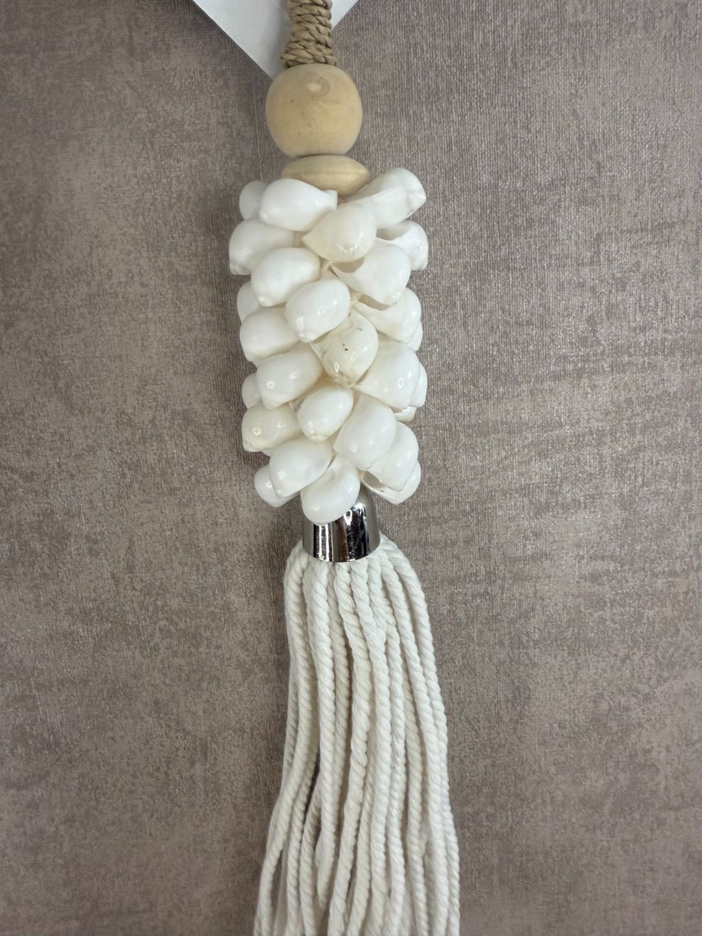 Witte hanger van schelpen en katoen met houten kraal en touw, hangend tegen een neutrale achtergrond.