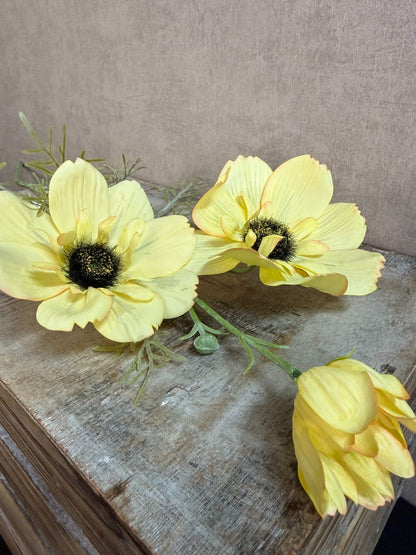 Gele kunstbloem anemoon met donkere harten en groene takjes tegen een neutrale achtergrond.