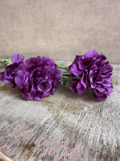 Paarse kunstbloem anjer met volle gelaagde bloemblaadjes, liggend op een houten ondergrond.