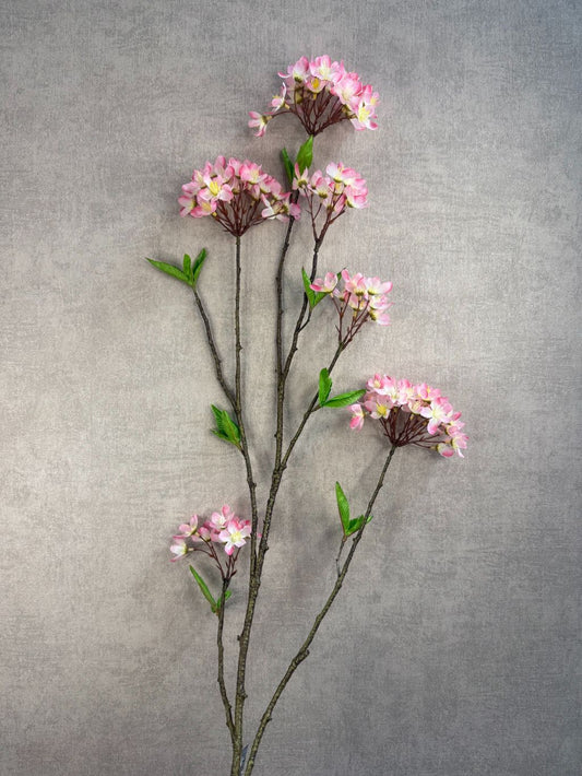 Lange kunsttak met vijf trosjes roze bloesem en groene blaadjes, tegen een grijze achtergrond.