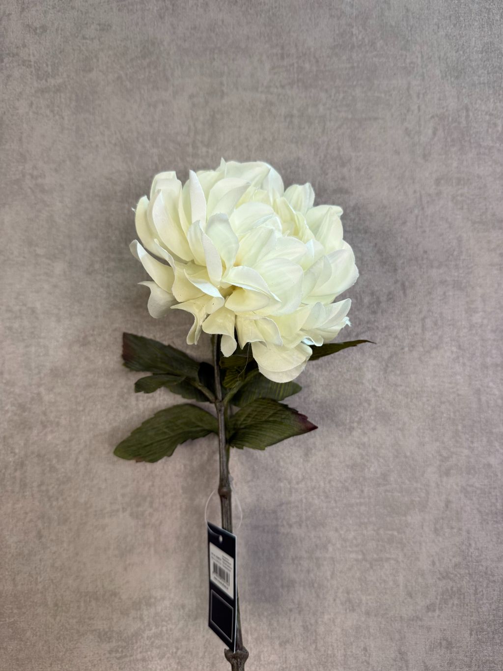 Crèmekleurige kunstbloem dahlia met volle bloem en groene bladeren tegen een neutrale achtergrond.