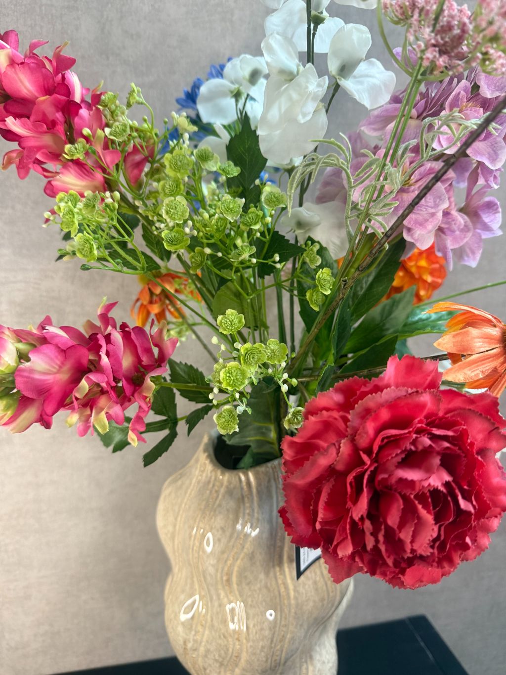 Kunstboeket met gemengde bloemen in diverse kleuren, geplaatst in een golvende beige vaas op zwarte ondergrond.