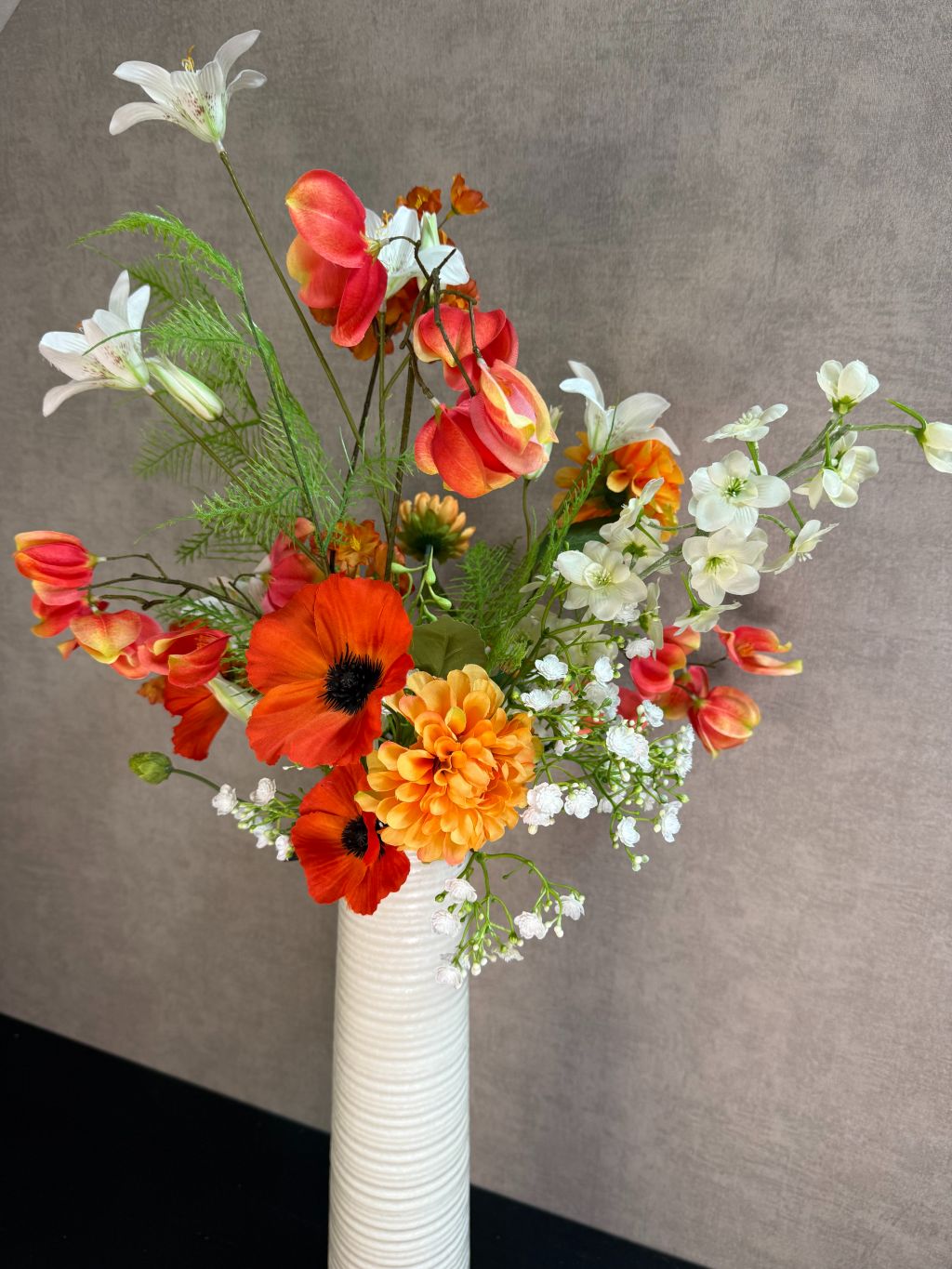 Kunstboeket met oranje en witte bloemen in een hoge crème vaas, geplaatst op een zwarte tafel tegen een neutrale achtergrond.