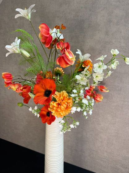 Kunstboeket met oranje en witte bloemen in een hoge crème vaas, geplaatst op een zwarte tafel tegen een neutrale achtergrond.