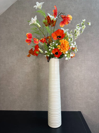 Kunstboeket met oranje en witte bloemen in een hoge crème vaas, geplaatst op een zwarte tafel tegen een neutrale achtergrond.