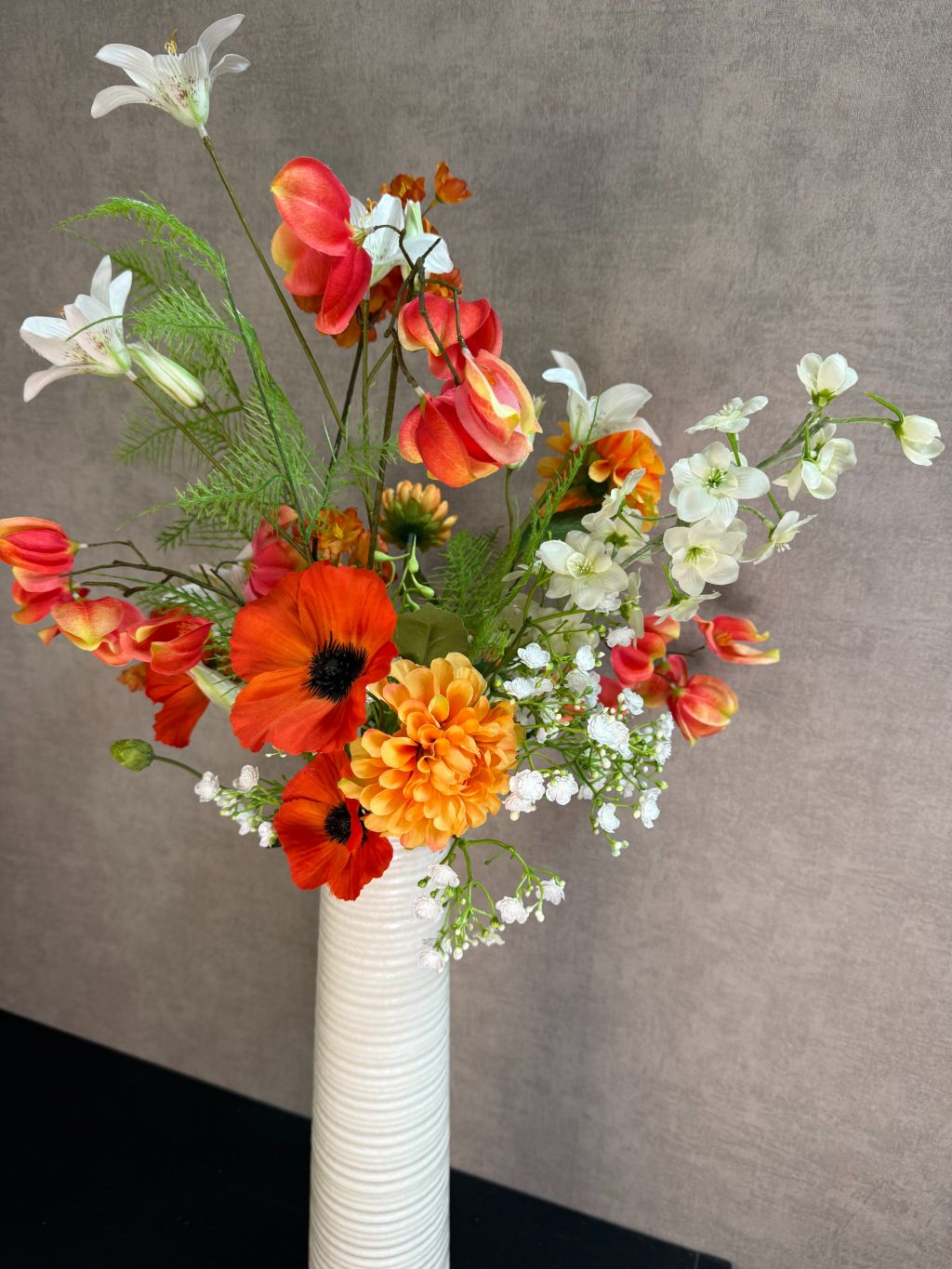 Kunstboeket met oranje en witte bloemen in een hoge crème vaas, geplaatst op een zwarte tafel tegen een neutrale achtergrond.