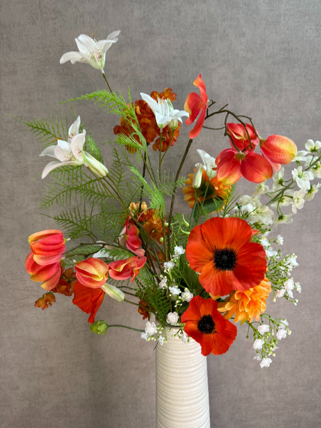 Kunstboeket met oranje en witte bloemen in een hoge crème vaas, geplaatst op een zwarte tafel tegen een neutrale achtergrond.