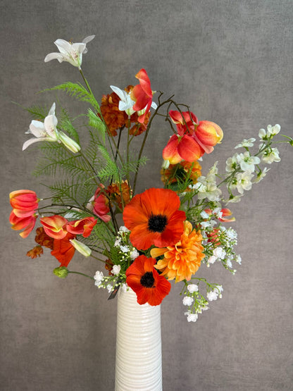 Kunstboeket met oranje en witte bloemen in een hoge crème vaas, geplaatst op een zwarte tafel tegen een neutrale achtergrond.