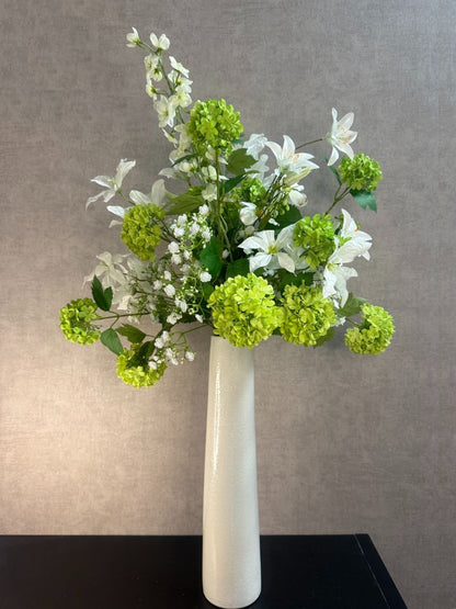 Kunstboeket met witte bloemen en groene sneeuwballen in een hoge crème vaas op zwarte ondergrond.