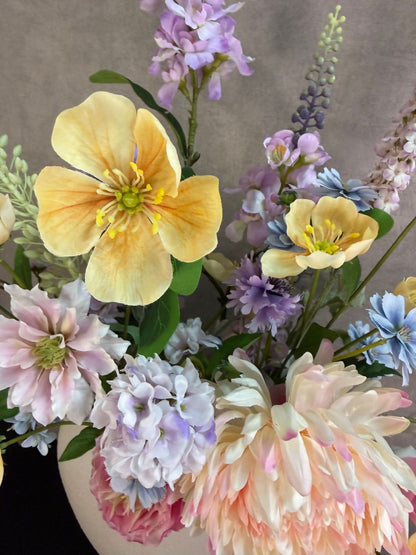 Groot pastelkleurig kunstboeket in een ronde witte vaas op een donkere ondergrond.