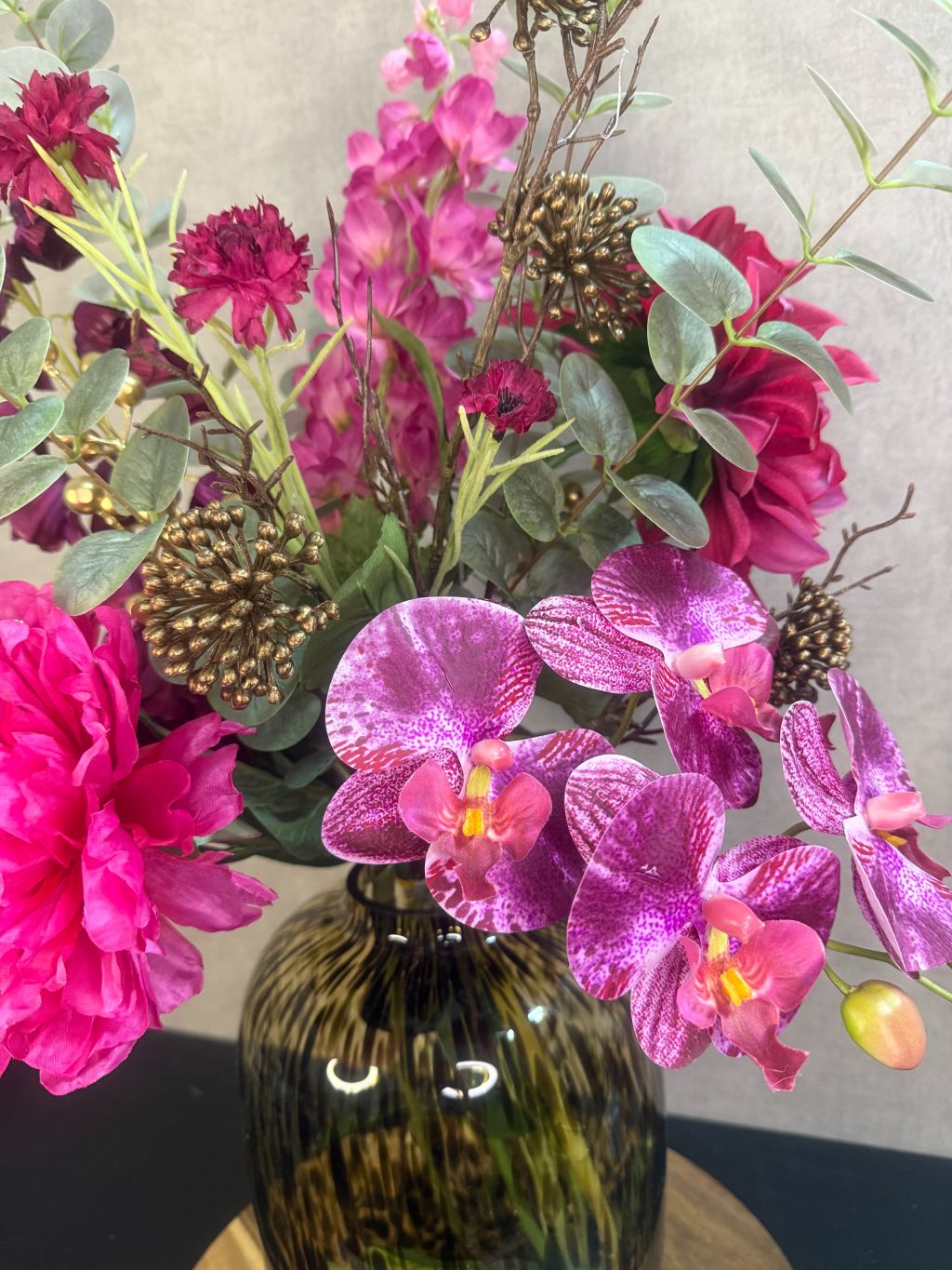 Kunstboeket met fuchsia pioenrozen, orchideeën en bordeaux bloemen in een glasvaas op houten ondergrond.