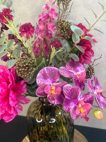 Kunstboeket met fuchsia pioenrozen, orchideeën en bordeaux bloemen in een glasvaas op houten ondergrond.