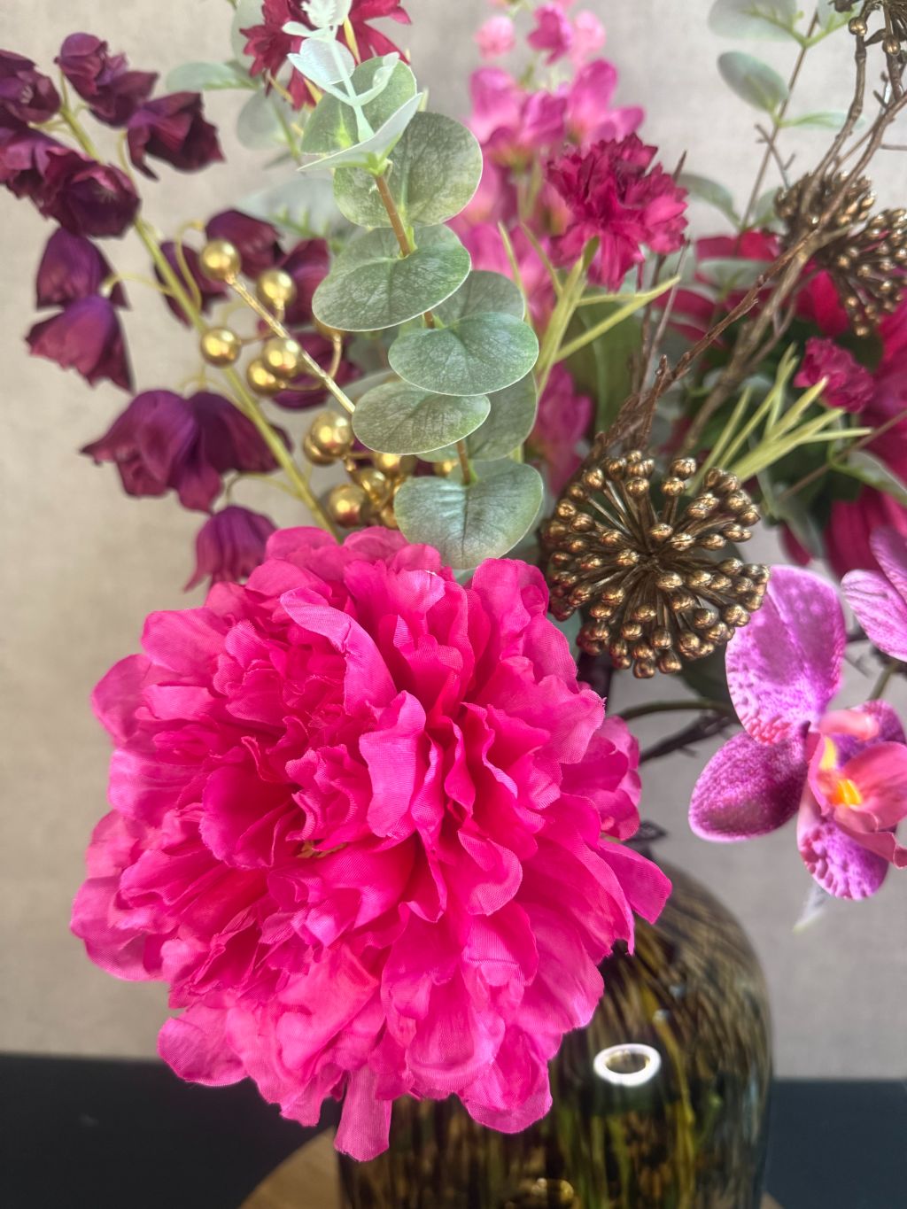 Kunstboeket met fuchsia pioenrozen, orchideeën en bordeaux bloemen in een glasvaas op houten ondergrond.