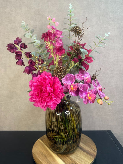 Kunstboeket met fuchsia pioenrozen, orchideeën en bordeaux bloemen in een glasvaas op houten ondergrond.