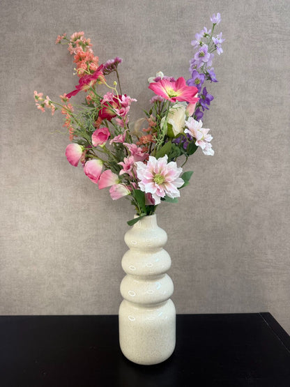 Kunstboeket in bonte kleuren met roze, lila en perzikbloemen in crèmekleurige vaas tegen grijze achtergrond.