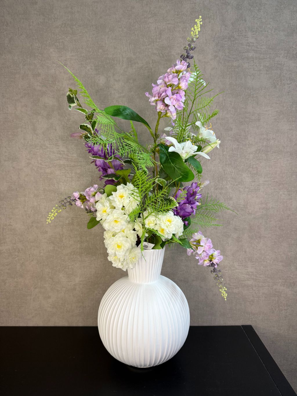 Paars en crèmekleurig kunstboeket met groene takken en witte bloemen in een witte ribbelvaas op een donkere tafel.