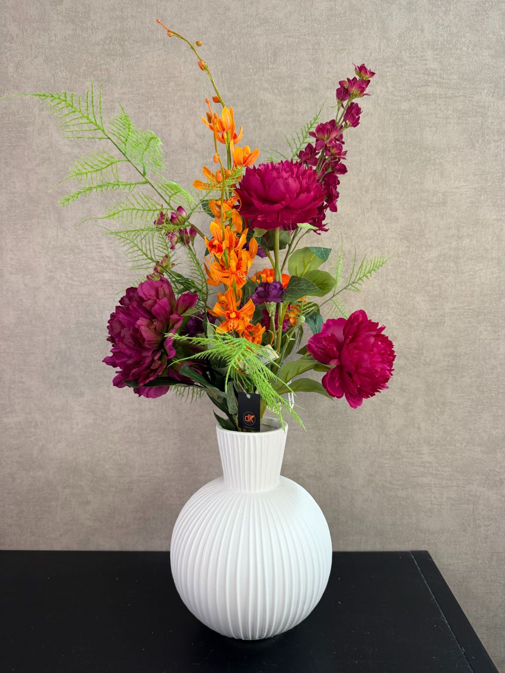 Kunstboeket met fuchsia, oranje en paarse bloemen in een witte vaas op een tafel tegen een neutrale achtergrond.