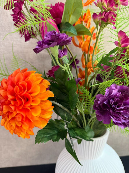 Kunstboeket met fuchsia, oranje en paarse bloemen in een witte vaas op een tafel tegen een neutrale achtergrond.
