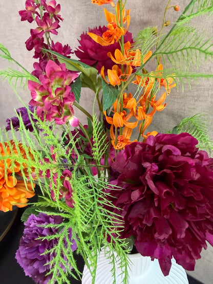 Kunstboeket met fuchsia, oranje en paarse bloemen in een witte vaas op een tafel tegen een neutrale achtergrond.