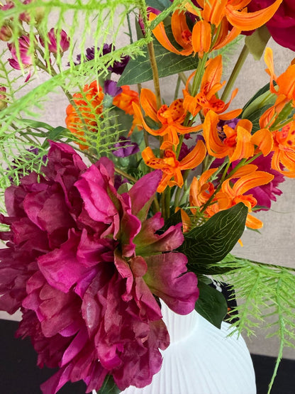 Kunstboeket met fuchsia, oranje en paarse bloemen in een witte vaas op een tafel tegen een neutrale achtergrond.