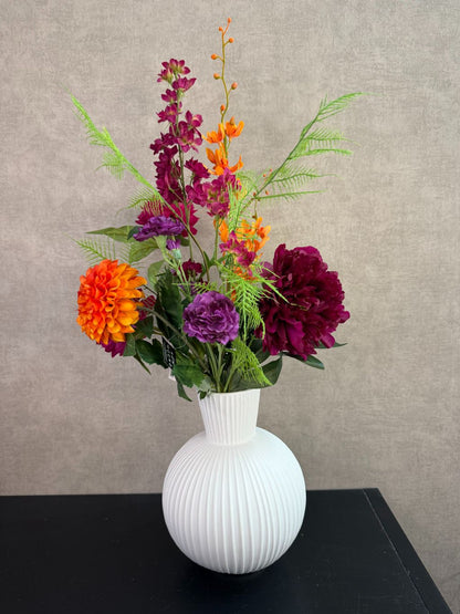 Kunstboeket met fuchsia, oranje en paarse bloemen in een witte vaas op een tafel tegen een neutrale achtergrond.