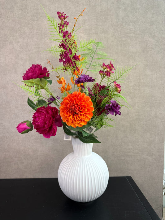 Kunstboeket met fuchsia, oranje en paarse bloemen in een witte vaas op een tafel tegen een neutrale achtergrond.