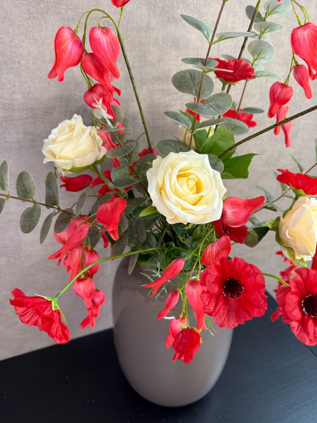 Kunstboeket met rode en crème bloemen in een grijze vaas op een tafel tegen een neutrale achtergrond.