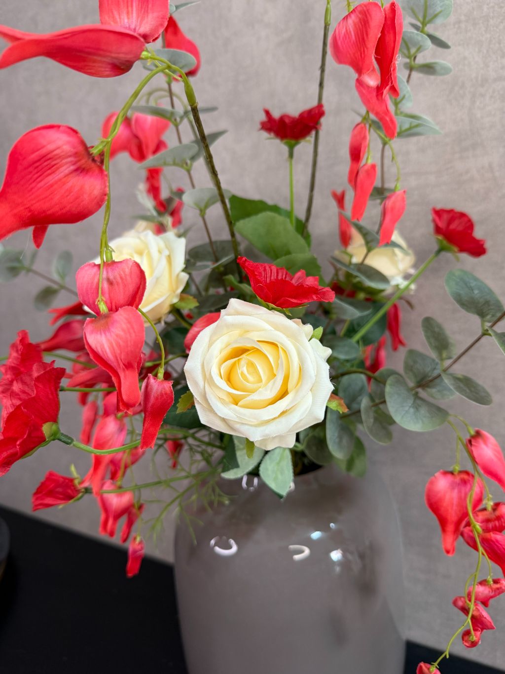 Kunstboeket met rode en crème bloemen in een grijze vaas op een tafel tegen een neutrale achtergrond.