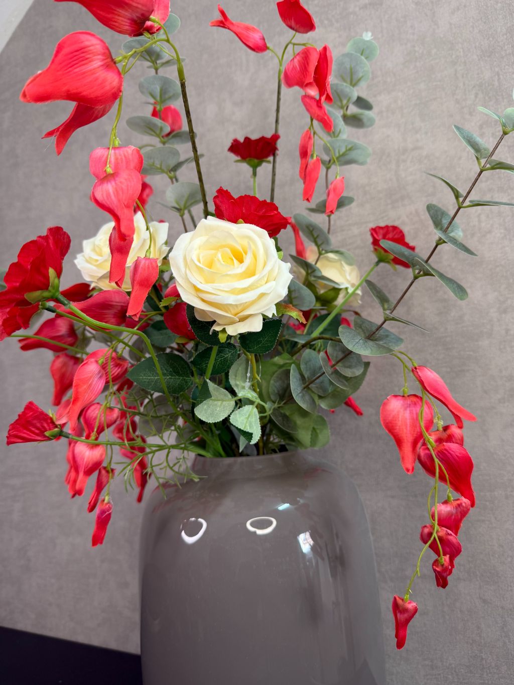 Kunstboeket met rode en crème bloemen in een grijze vaas op een tafel tegen een neutrale achtergrond.