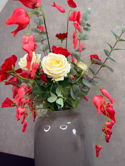 Kunstboeket met rode en crème bloemen in een grijze vaas op een tafel tegen een neutrale achtergrond.