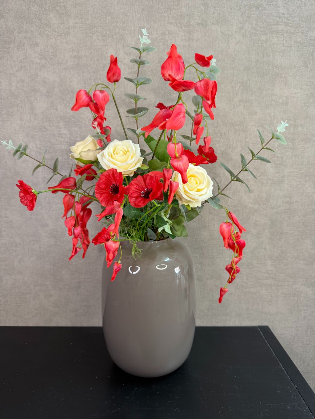 Kunstboeket met rode en crème bloemen in een grijze vaas op een tafel tegen een neutrale achtergrond.