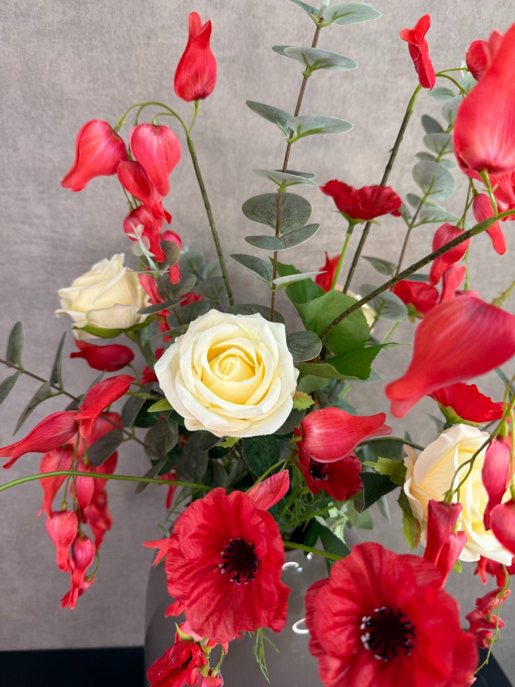 Kunstboeket met rode en crème bloemen in een grijze vaas op een tafel tegen een neutrale achtergrond.