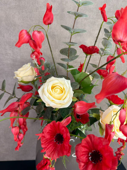 Kunstboeket met rode en crème bloemen in een grijze vaas op een tafel tegen een neutrale achtergrond.
