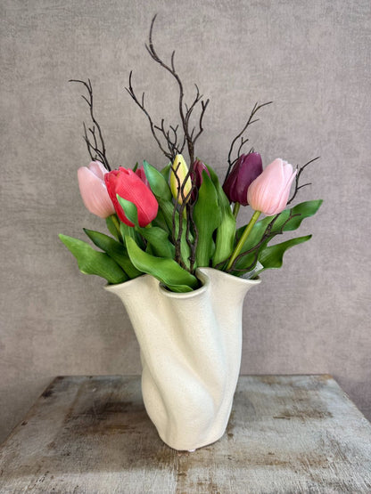 Kunstboeket met paars- en rozekleurige tulpen en groene bladeren in een lichte vaas op een houten ondergrond.