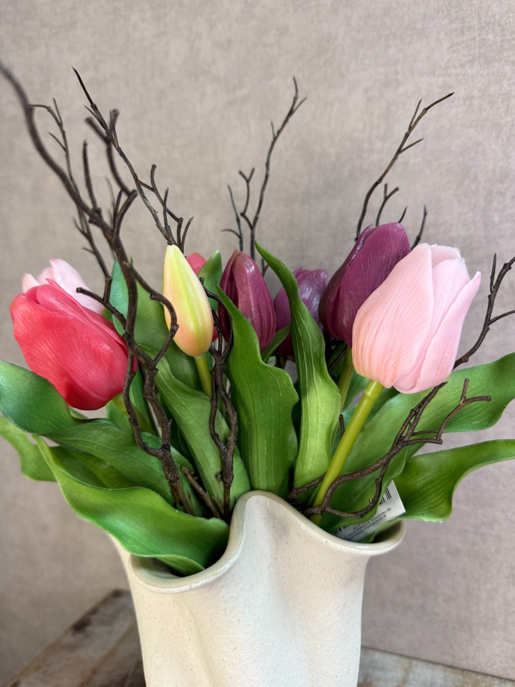 Kunstboeket met paars- en rozekleurige tulpen en groene bladeren in een lichte vaas op een houten ondergrond.