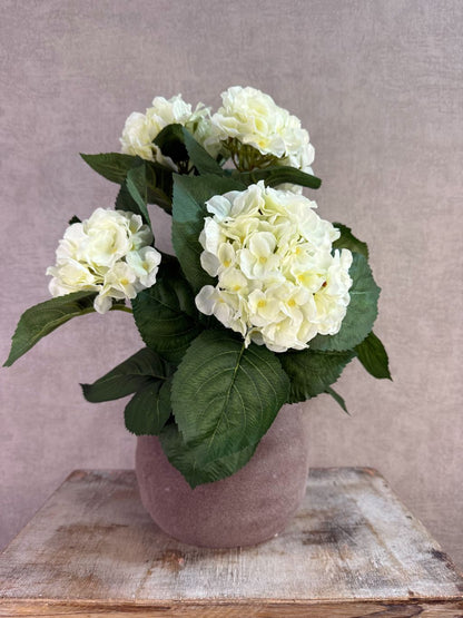 Witte kunst hortensia met groene bladeren in een bruine pot op een houten ondergrond.