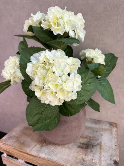 Witte kunst hortensia met groene bladeren in een bruine pot op een houten ondergrond.